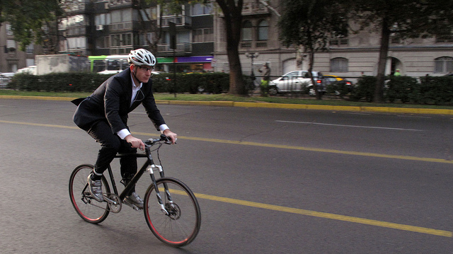 Homem de terno e capacete, pedalando uma bicicleta preta em dire